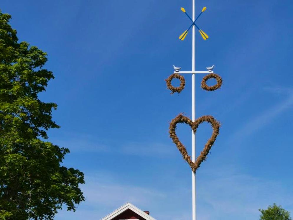 Mittsommerfest in Schweden: Blüten, Schnaps und lange Nächte. Jeder Ort schmückt seinen Midsommarbaum nach eigenen Traditionen und mit eigenen Symbolen – bei dieser Stange vor einer Festhalle in Rättvik sieht man etwa ein Herz, Ringe sowie Tauben. 