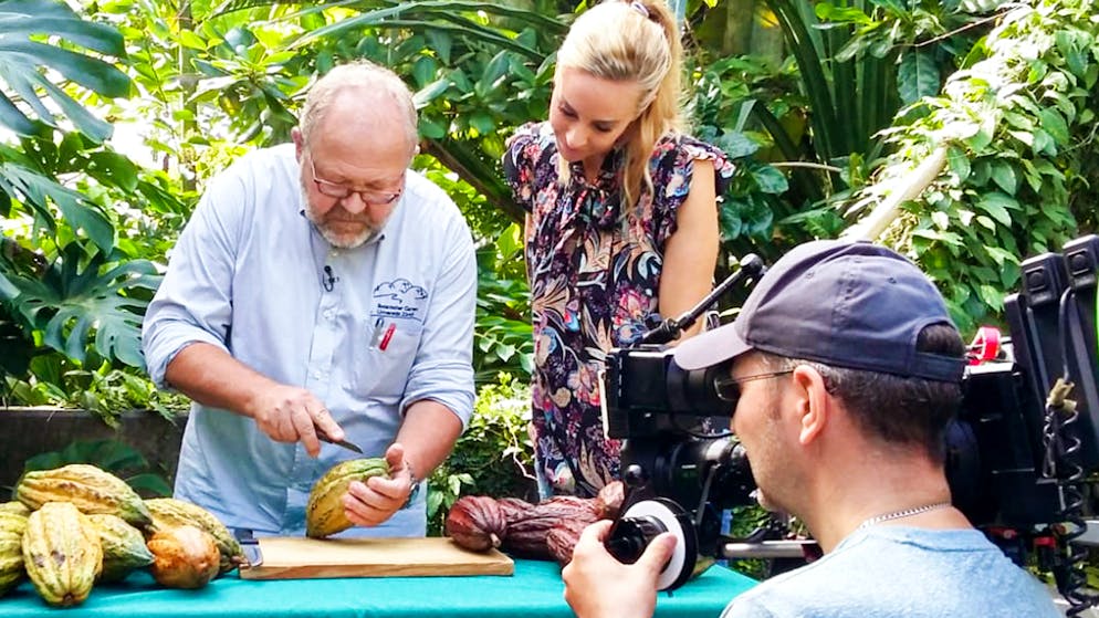 Nicole Berchtold in «Was kostet die Schweiz»?. Im botanischen Garten der Universität Zürich zeigt ein Mitarbeiter Nicole Berchtold, wie die Kakaofrüchte aussehen und schmecken.