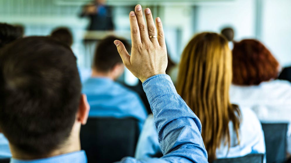 Rear view of a businessman raising his hand to ask the question on a seminar with large group of his colleagues. Focus is on hand.