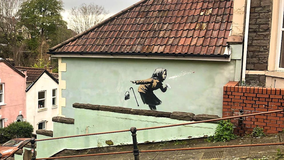 Banksy und Corona. Banksy lässt eine ältere Frau auf einer Wand in der Vale Street in Bristol die benachbarten Häuser wegniesen.
