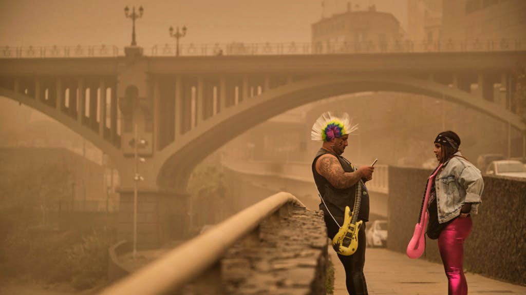 Atmosfera pesante alle Canarie: due persone in costume di carnevale si intrattengono lungo un marciapiede di Santa Cruz de Tenerife avvolti da un’aria intrisa di polvere rossa. Il traffico aereo e le celebrazioni del carnevale nelle isole Canarie sono stati gravemente colpiti da una tempesta di sabbia e violente raffiche.