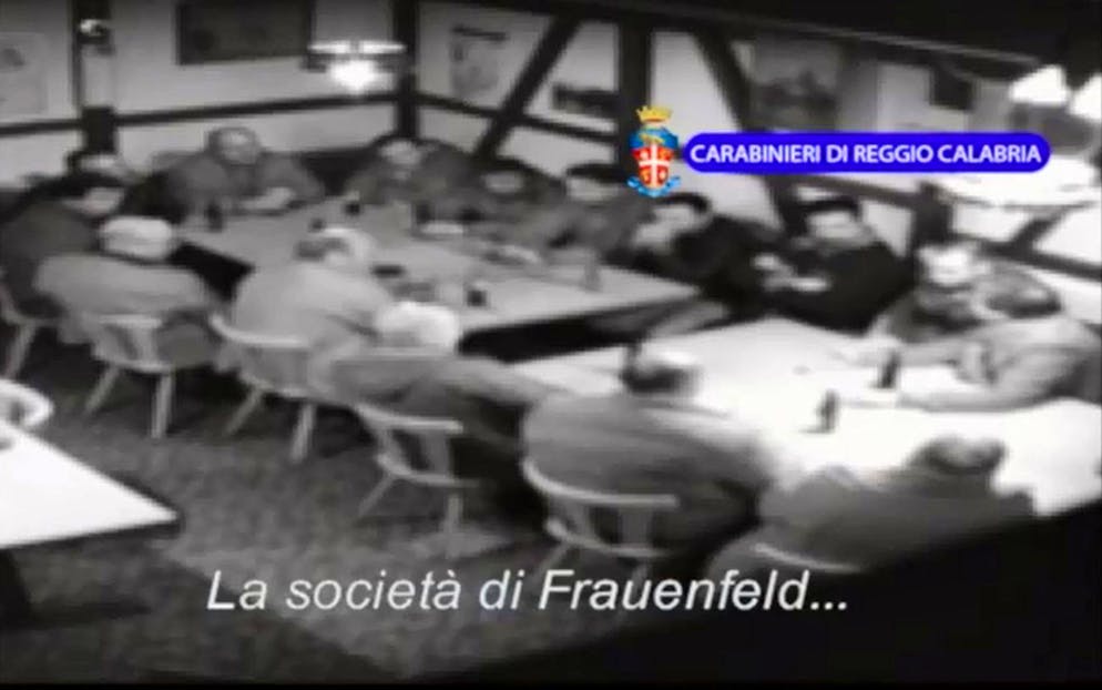 Nell'immagine d'illustrazione un frammento della sequenza video dell''«Operazione Helvetica», che la polizia calabrese ha messo in rete venerdì 22 agosto 2014. Nella foto si vedono una dozzina di uomini attorno a due tavoli, probabilmente in un ristorante. Le conversazioni sbiadite in accento del sud Italia ruotano attorno alle regole in vigore dal 1830, attorno a opportunità di affari come ricatti, cocaina, eroina e sulla «Società a Frauenfeld». Seguirono 15 arresti di uomini dell'Ndrangheta in un'azione coordinata nei cantoni di Turgovia, Zurigo e Vallese.