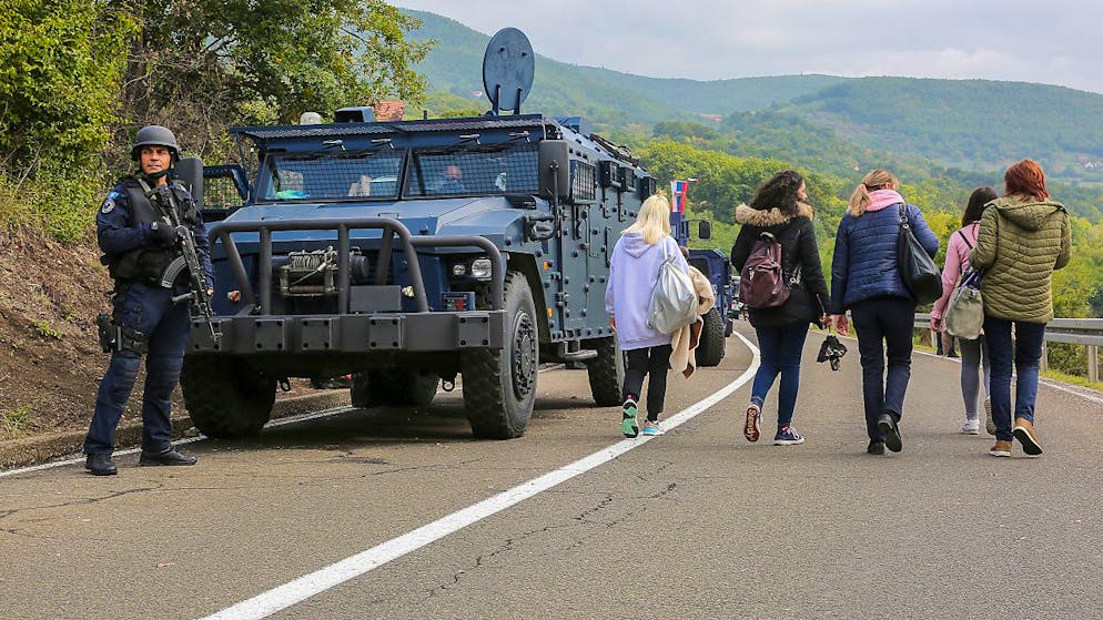 Ein kosovarischer Soldat hält eine Waffe in der Hand und steht auf einer Straße in der Nähe des Grenzortes Jarinje, während Personen an ihm vorbeigehen. Zwischen dem Kosovo und Serbien ist es wegen einer neuen Verordnung über die Zulässigkeit von Kfz-Kennzeichen zu Spannungen gekommen. Foto: Visar Kryeziu/AP/dpa