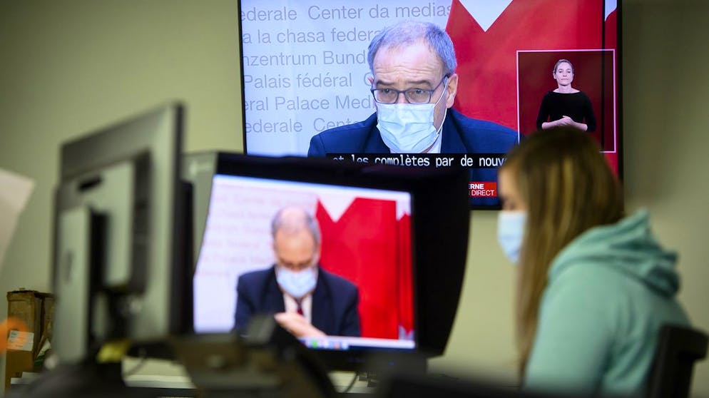 A journalist wearing face mask watchs Swiss President Guy Parmelin speaking during a press conference in Bern on TV screens inside an office announcing new measures against the Coronavirus (COVID-19) in Lausanne, Switzerland, Wednesday, January 13, 2021. (KEYSTONE/Laurent Gillieron)