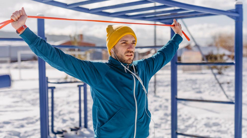Young man with knitted cap with resistance bands doing exercises outdoor on snow