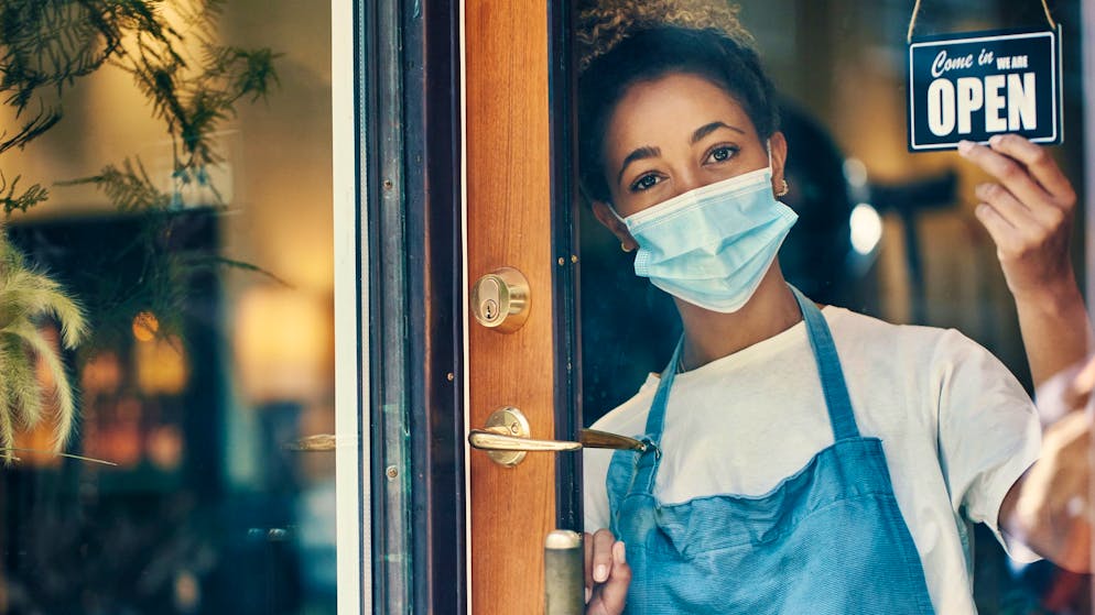 Shot of a masked young woman hanging an open sign on the window of a cafe