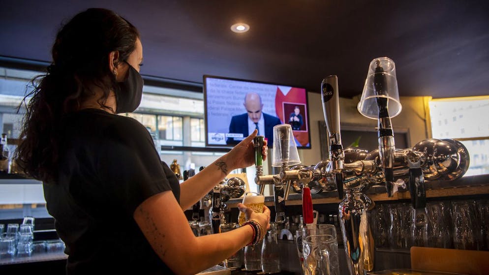 A waitress is preparing a beer wearing a protective mask while watching on a TV screen Swiss Interior and Health Minister Alain Berset as he speaks during a press conference in Bern announcing new measures against the Coronavirus (COVID-19), inside of the bar-restaurant