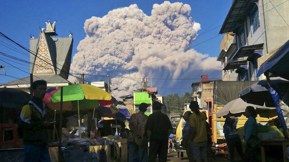 People watch as Mount Sinabung spews volcanic material during an eruption, at a market in Karo, North Sumatra, Indonesia, Tuesday, March 2, 2021. The 2,600-metre (8,530-feet) volcano erupted Tuesday, sending volcanic materials a few thousand meters into the sky and depositing ash on nearby villages. (AP Photo/Sugeng Nuryono)