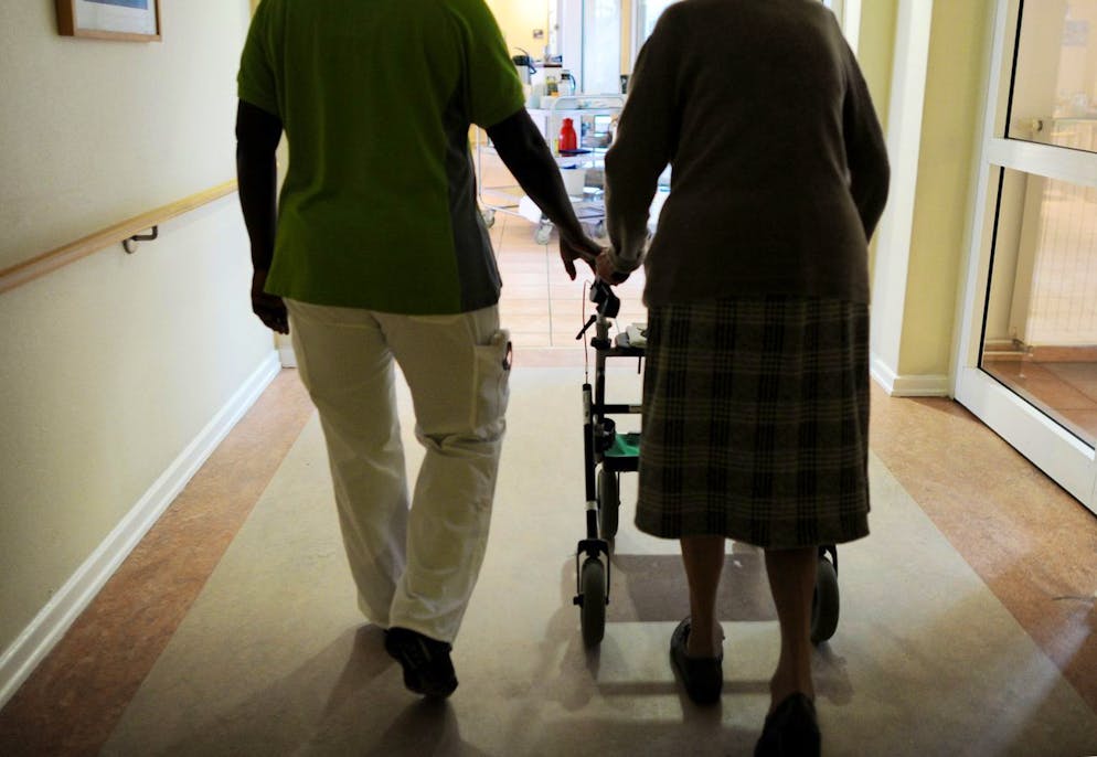ARCHIV - Eine Pflegehausbewohnerin und eine Pflegehelferin gehen am 08.03.2013 in Hamburg in einem Seniorenzentrum mit einem Rollator einen Gang entlang. Die Grünen haben Union und SPD aufgefordert, die Reform der Pflegeausbildung nicht wegen Streitereien innerhalb der großen Koalition auf die lange Bank zu schieben. (zu dpa