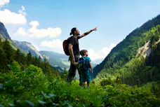 Ein Vater zeigt seinem Kind beim Wandern in einem grünen Bergthal den Ausblick.