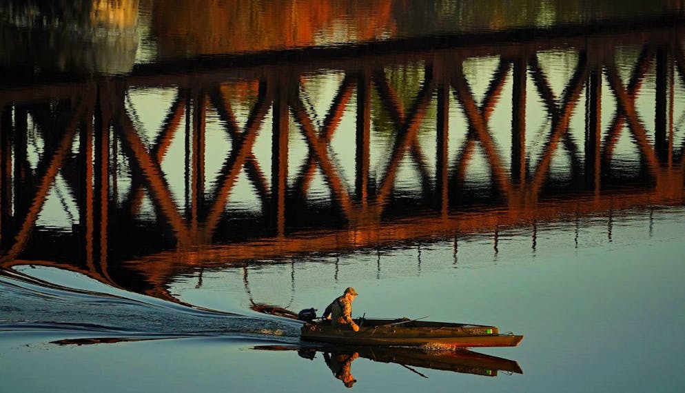 A duck hunter motors by the reflection of a train trestle on the Androscoggin River, Friday, Oct. 29, 2021, in Brunswick, Maine. (AP Photo/Robert F. Bukaty)