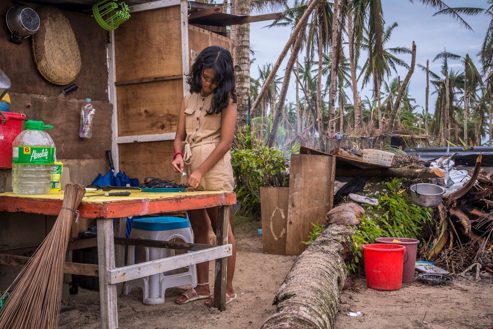 Liza May Antipasado, 19 years old in brown dress while cooking. Liza is preparing Torta Talong (burned eggplant) for the whole family of 6 in a makeshift kitchen underneath a fallen coconut tree