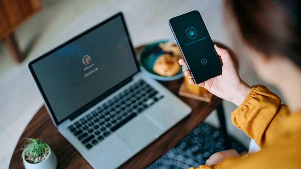 Young Asian woman logging in to her laptop and holding smartphone on hand with a security key lock icon on the screen, sitting in the living room at cozy home. Privacy protection, internet and mobile security concept
