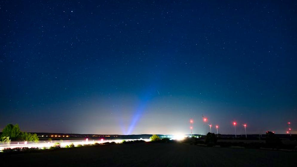 PRODUKTION - 06.05.2022, Brandenburg, Jacobsdorf: Hell erleuchtet ist die Autobahn A12, am Rand das rote Licht der Windenergieanlagen und im Hintergrund erhellt blaues Licht den Nachthimmel über der Stadt Frankfurt (Oder). Angestrahlte Kirchen und Denkmäler, Flutlicht im Stadien und grelle Straßenbeleuchtung - die Menschen machen vielerorts die Nacht zum Tag, mit verheerenden Folgen für die Tierwelt und die Artenvielfalt. (zu dpa-KORR.: