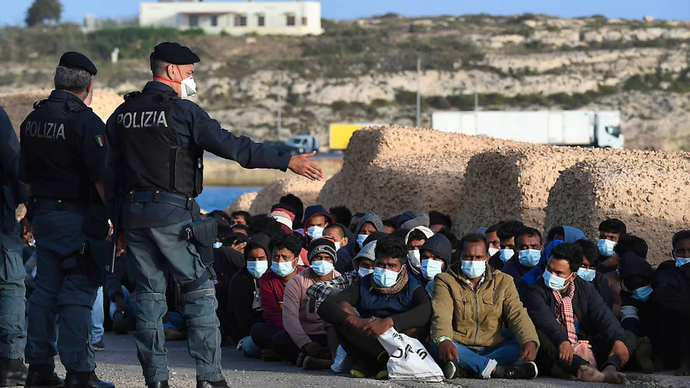 dpatopbilder - ARCHIV - Italienische Polizisten (l) stehen auf der kleinen italienischen Mittelmeerinsel Lampedusa neben Migranten, die Mund-Nasen-Schutz tragen und auf dem Boden sitzen. Foto: Salvatore Cavalli/AP/dpa
