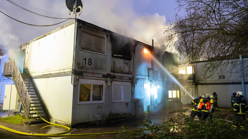 Feuerwehrleute stehen bei Löscharbeiten vor den verqualmten und teils brennenden Container-Einheiten einer Flüchtlingsunterkunft in Hamburg-Winterhude.