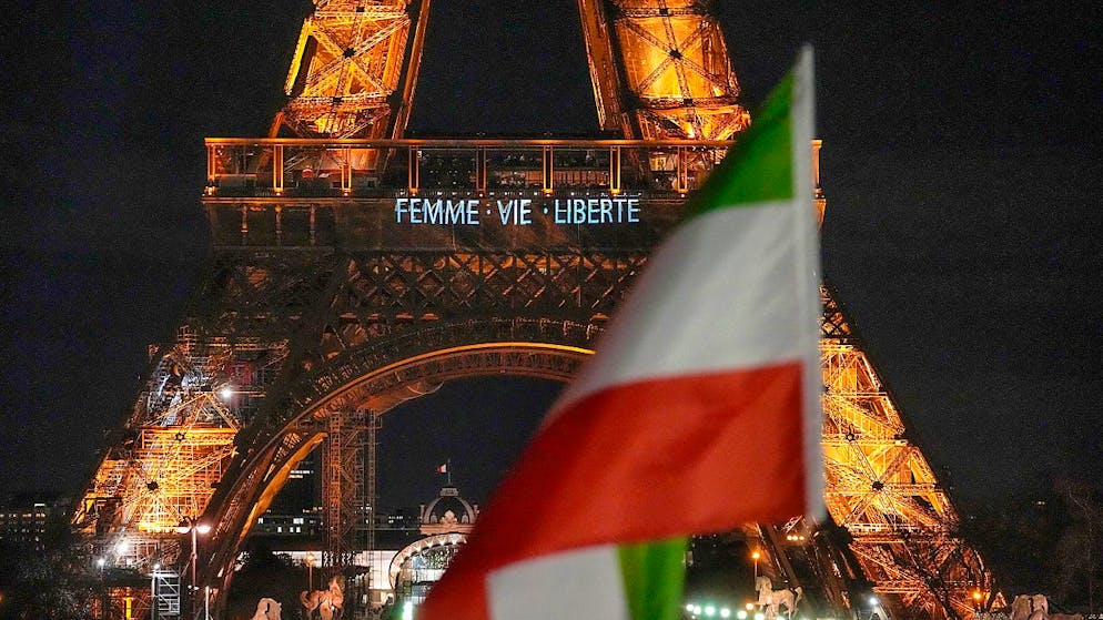 dpatopbilder - ARCHIV - Der Eiffelturm leuchtet in der Nacht mit dem Slogan «Frau, Leben, Freiheit» zur Unterstützung der Proteste im Iran, während im Vordergrund eine iranische Fahne weht. Foto: Michel Euler/AP/dpa