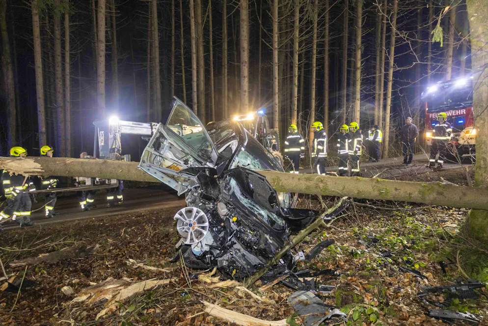 Feuerwehrleute betrachten das Auto im österreichischen Aurolzmünster, das in einen liegenden Baumstamm krachte.