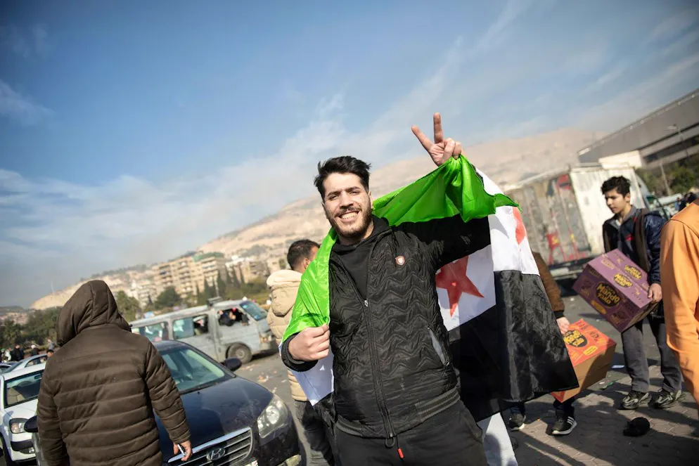 Der Umsturz in Syrien in Bildern. Ein Mann feiert den Umsturz in Damaskus eingehüllt in eine revolutionäre Flagge. Diese zeigt auf grün-weiss-schwarzem Grund drei rote Sterne im Gegensatz zur – bislang – offiziellen rot-weiss-schwarzen Landesfahne mit zwei grünen Sternen, die als Symbol der Langzeitherrschaft des Assad-Clans im ganzen Land verhasst ist.