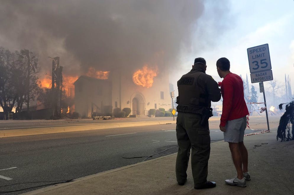Inferno di fuoco a Los Angeles, migliaia di persona in fuga. Il vice dello sceriffo di Los Angeles, a sinistra, parla con un residente mentre la chiesa della comunità di Altadena brucia sullo sfondo.