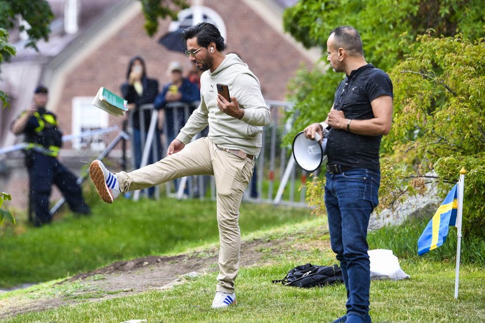 Salwan Momika tritt am 20. Juli 2023 eine Koran-Ausgabe. Für Muslime ist der Koran heilig: Die Aktion sorgte für viel Aufruhr. Rechts neben ihm sieht ein Polizist zu, der mit einem Megafon bewaffnet ist.