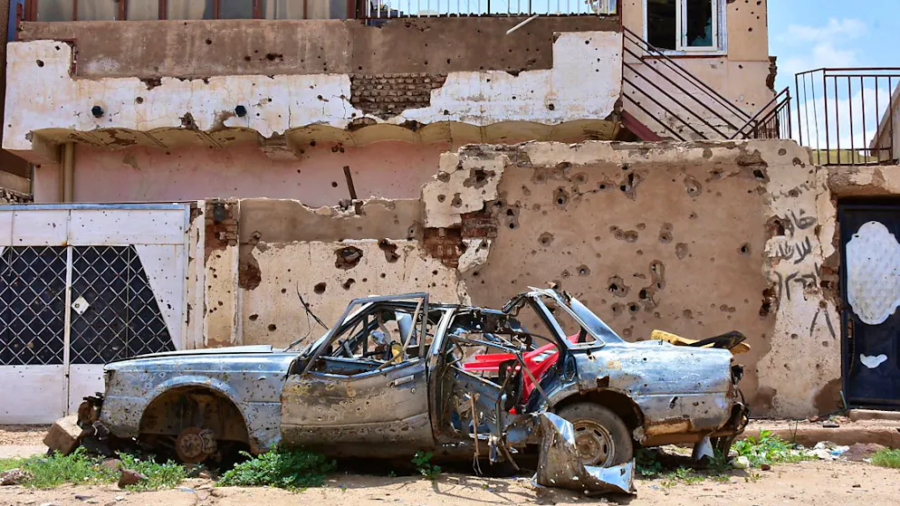 ARCHIV - Ein zerstörtes Auto steht vor einem Haus voller Einschüsse in Omdurman, Sudan. Foto: Mudathir Hameed/dpa/Archivbild