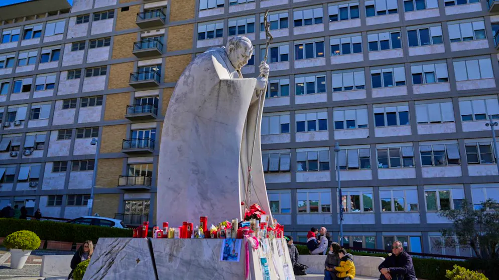 Menschen sitzen bei der Statue von Papst Johannes Paul II. auf dem Gelände der Poliklinik Agostino Gemelli, wo Papst Franziskus seit Mitte Februar behandelt wird. Foto: Gregorio Borgia/AP/dpa