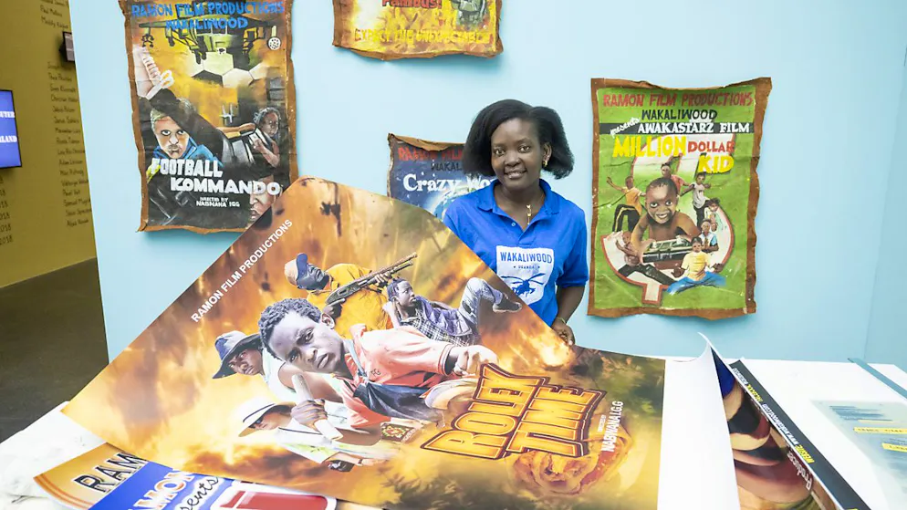 Kunstmuseum Luzern zeigt Collagen von Małgorzata Mirga-Tas - Gallery. Die Co-Produzentin von Wakaliga, Harriet Nakasujja, präsentiert das Plakat zum Film "Rolex Time". Im Hintergrund gemalte Plakate zu früheren Wakaliga-Filmen.