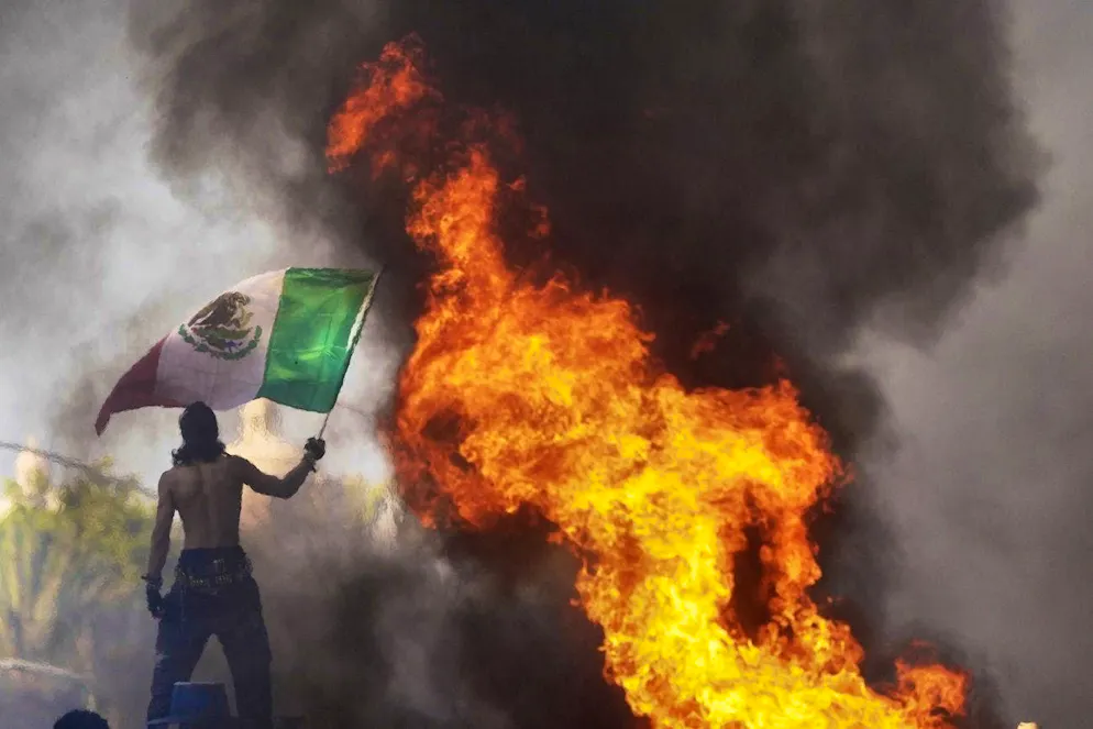 Protests L.A.. A demonstrator waves a Mexican flag as a Waymo cab burns near the Metropolitan Detention Center in downtown Los Angeles.