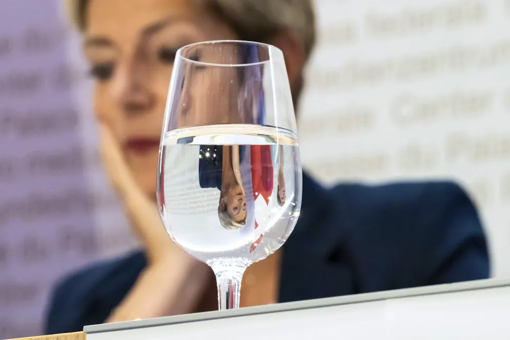 La présidente de la Confédération Karin Keller-Sutter se reflète dans un verre d'eau lors d'une conférence de presse sur le paquet d'allègements fiscaux 27 et le budget 2026, le mercredi 25 juin 2025, au Centre de presse du Palais fédéral à Berne. (KEYSTONE/Anthony Anex)