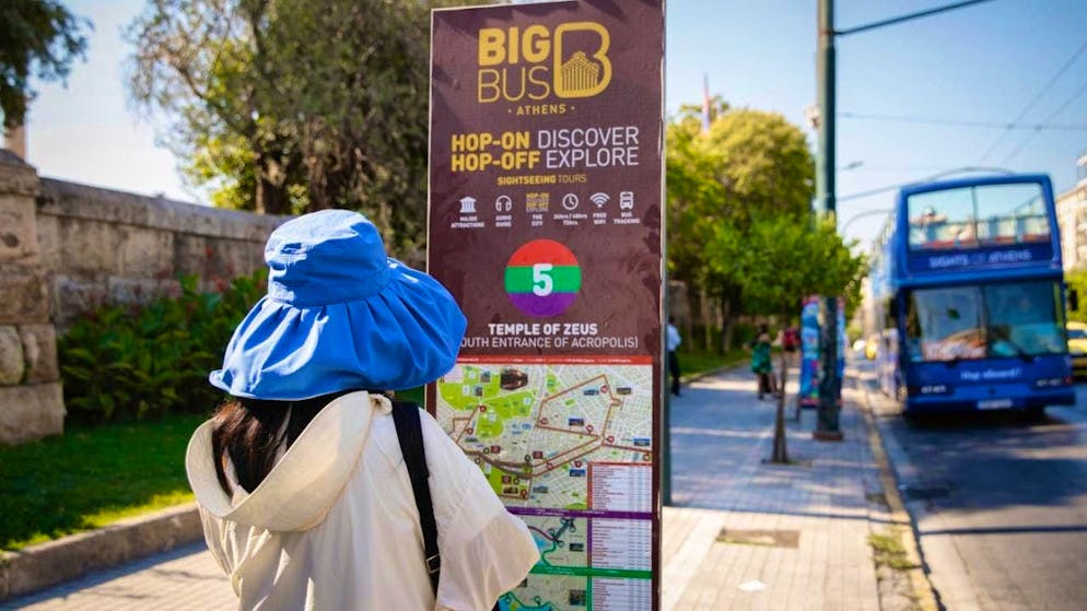 A tourist in a sun hat stands at a sightseeing bus stop near the Acropolis. Photo: Socrates Baltagiannis/dpa