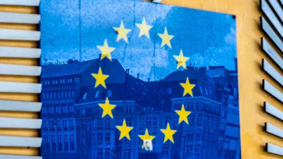 PRODUKTION - Drei Fahnen der Europäischen Union und gegenüberliegende Gebäude spiegeln sich im EU-Logo an der Außenfassade des Berlaymont-Gebäudes. Foto: Alicia Windzio/dpa