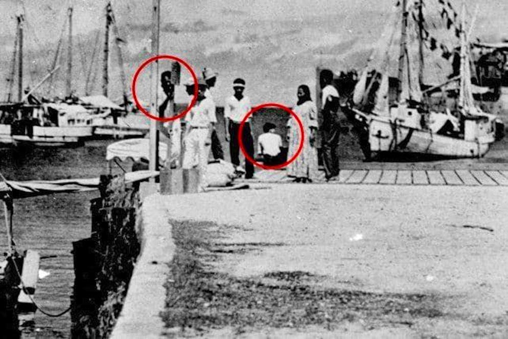 Amelia Earhart in immagini. Dettaglio ingrandito della foto precedente: mostra una donna seduta con i capelli ricci, che si dice assomigli ad Amelia Earhart, e una figura che potrebbe essere il suo navigatore Fred Noonan. Sul lato destro dell'immagine è visibile un oggetto sfocato, che alcuni interpretano come parte del relitto del suo Lockheed Electra.