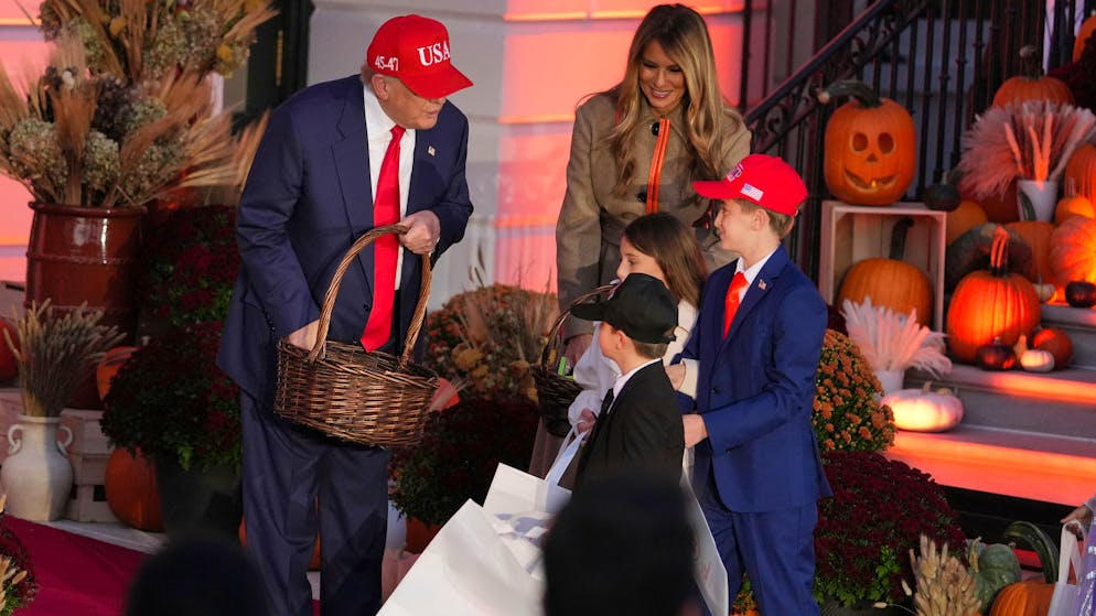 US-Präsident Donald Trump (l.) – in seiner üblichen Kluft – und First Lady Melania Trump – immerhin in einem mit der Halloween-Farbe Orange verzierten Trenchcoat – verteilen Süssigkeiten an Kinder, die als First Lady, Trump und Secret-Service-Mitglied verkleidet sind. (30. Oktober 2025)