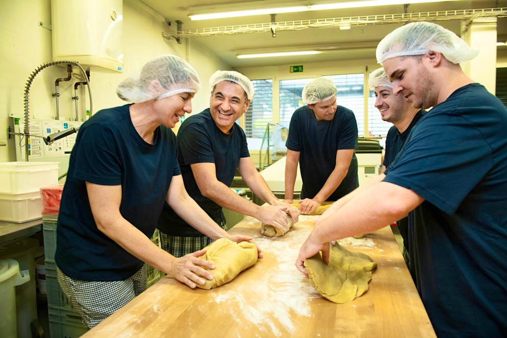 UBS Helpetica cuisson. Travail d’équipe dans la boulangerie: Nicole, Umut, Vincent, Davide et Tobias pétrissent la pâte ensemble. Umut et Vincent sont des collaborateurs de la boulangerie, Nicole, Davide et Tobias sont bénévoles.