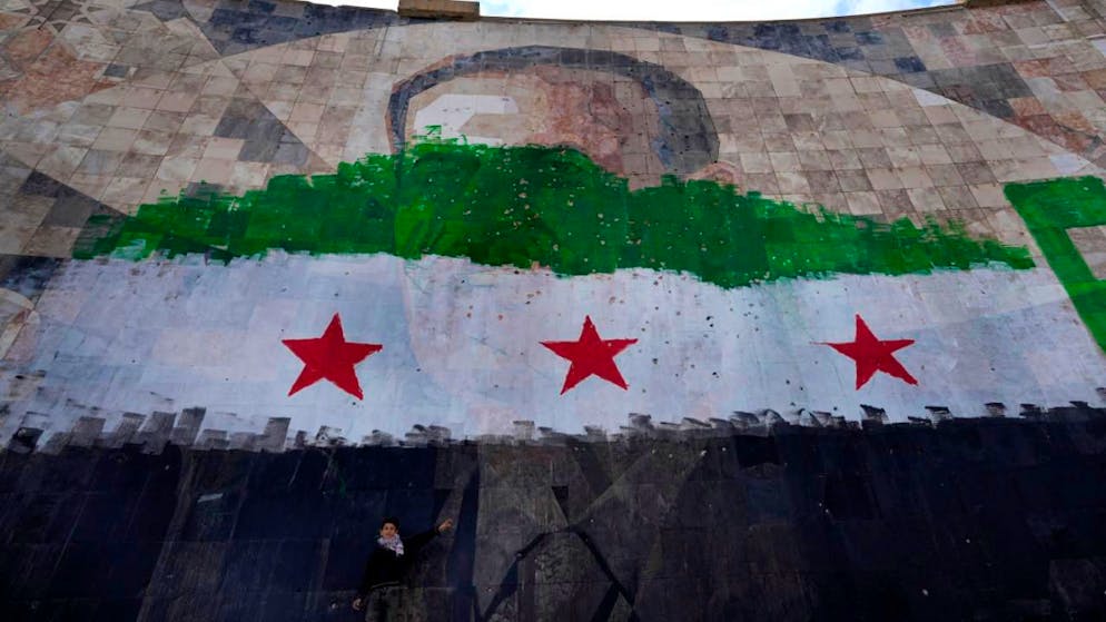 ARCHIV - Ein syrischer Junge steht unter einem riesigen Porträt des verstorbenen syrischen Präsidenten Al-Assad, übermalt mit den Farben der sogenannten "revolutionären" Flagge, in Damaskus. Foto: Hussein Malla/AP/dpa/Symbolbild