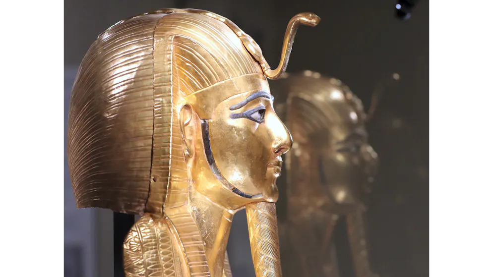 Psusennes_Tanis. Die lebensgrosse Goldmaske des Psusennes I. mit ruhigen, idealisierten Gesichtszügen: Sie trägt das gestreifte Nemes-Kopftuch, auf der Stirn sitzt die Uräus-Kobra als königliches Schutzsymbol. Auffällig sind die dunkel konturierten Augen und Details in blau – sie entstehen durch Einlagen aus Lapislazuli und Glasfluss. Am Kinn sitzt der geflochtene Zeremonialbart, ein typisches Zeichen königlicher Würde.