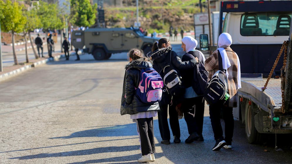Palästinensische Studenten warten am Strassenrand, während israelische Soldaten am 10. Dezember 2025 in der Nähe der Siedlung Kiryat Arba in Hebron mit einem gepanzerten Fahrzeug den Zugang blockieren.