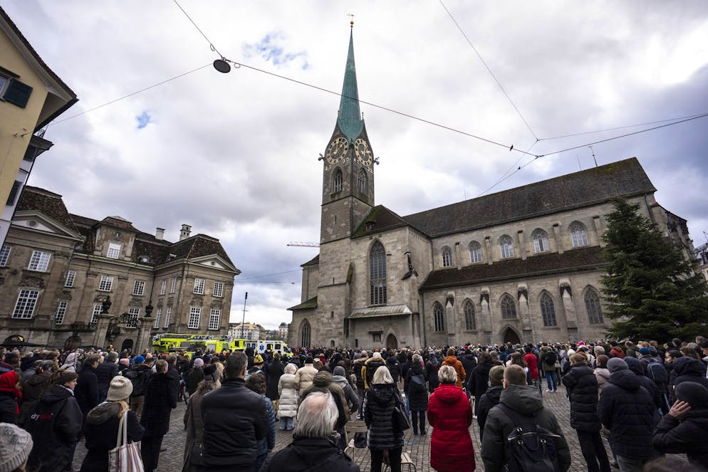 La Suisse pleure. Des personnes regardent la tour Fraumuenster pendant un hommage de 5 minutes marqué par le son des cloches lors de la cérémonie commémorative et de la journée nationale de deuil qui a suivi l'incendie mortel du bar « Le Constellation » à Crans-Montana, sur la Muensterplatz à Zurich, en Suisse, le vendredi 9 janvier 2026.