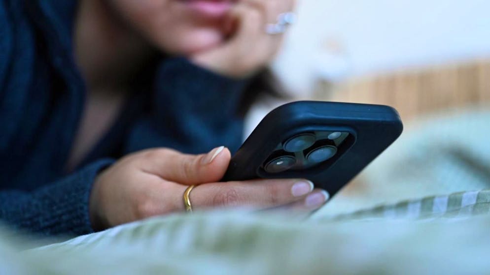 ILLUSTRATION - Eine junge Frau liegt mit dem Smartphone in der Hand auf dem Bett. Foto: Elisa Schu/dpa