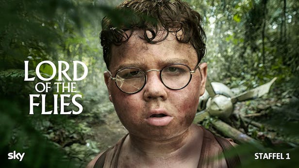 Nahaufnahme eines schmutzigen Jungen mit Brille, deren eines Glas zerbrochen ist, im Hintergrund ist ein Flugzeugwrack im dichten Dschungel zu erkennen.