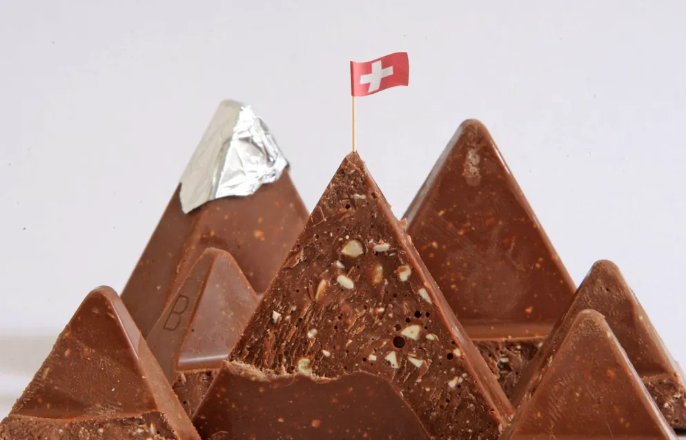 Schnitzelbängg 2026 Schweiz. Bitz weeniger steggt dinn zum glyche Bryys – au in dr Schwyz<br>Uf ere Toblerone – sait me – fäält ab jetz e Spitz.<br>Gisch aifach nit der Düürig tschuld am Bschiss in däm Regal!<br>Nai, unsri Bärge mööge nümm und stürze sich ins Daal.<br><br>(Schlachtbangg)