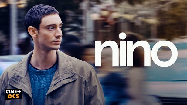 Ein junger Mann mit kurzen dunklen Haaren und einer grauen Jacke blickt nachdenklich zur Seite vor einem verschwommenen Stadthintergrund.