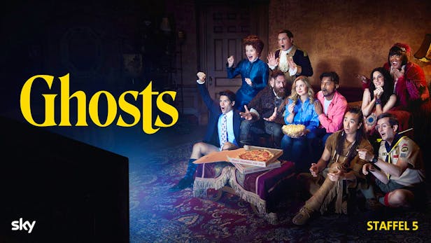 Eine Gruppe von Menschen und historisch gekleideten Geistern sitzt erschrocken auf einem Sofa vor einem Fernseher und isst Pizza und Popcorn.