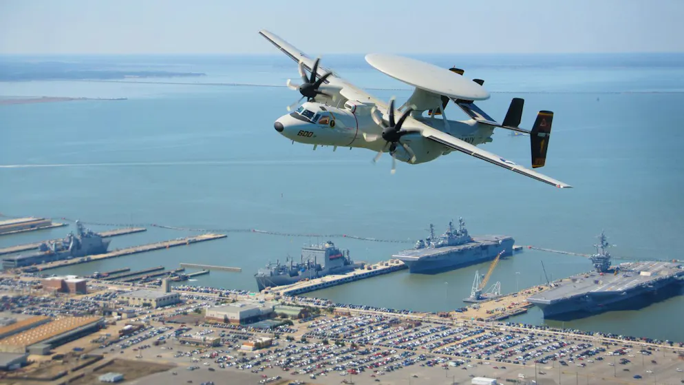 Eine E-2D Hawkeye fliegt über den Marinestützpunkt Norfolk in Virginia. Der Flugzeugträger USS Theodore Roosevelt ist auch zu sehen.