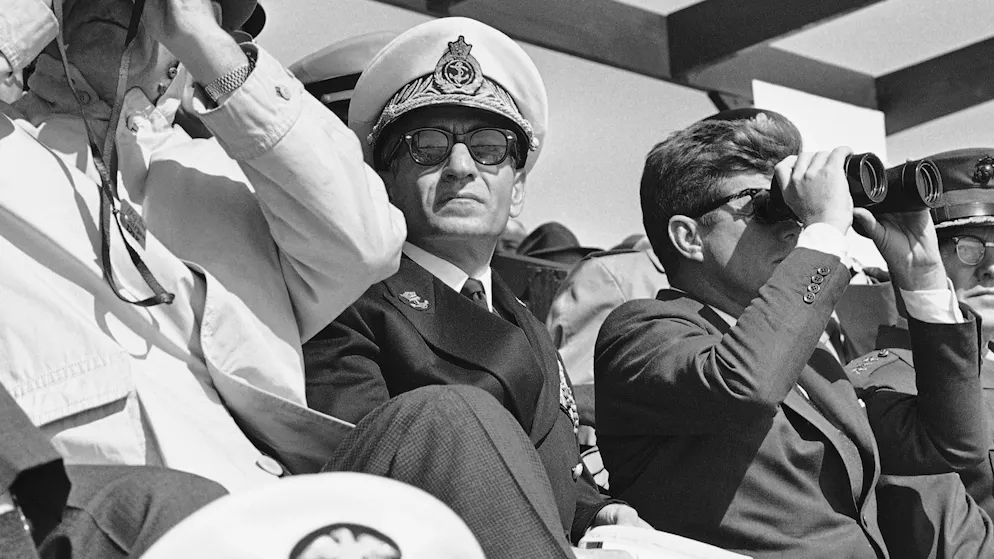 Der Schah des Iran, Mohammad Reza Pahlavi, sitzt im April 1962 zwischen Präsident John F. Kennedy und Vizepräsident Lyndon B. Johnson, während sie durch Ferngläser Marine-Landungsmanöver im Camp LeJeune in North Carolina beobachten.