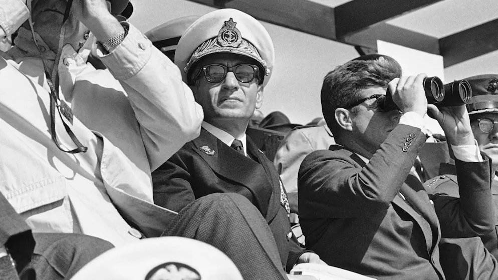 Le shah d'Iran, Mohammad Reza Pahlavi, est assis entre le président John F. Kennedy et le vice-président Lyndon B. Johnson en avril 1962, alors qu'ils observent à travers des jumelles des manœuvres de débarquement naval au camp LeJeune en Caroline du Nord.