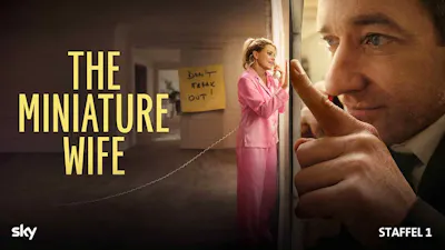 Ein Mann blickt mit riesigem Gesicht durch eine Türöffnung in einen Raum. Dort steht eine Frau in einem pinken Pyjama, die im Vergleich zu ihm winzig klein ist und telefoniert. An der Wand im Hintergrund klebt eine Notiz mit der Aufschrift: 