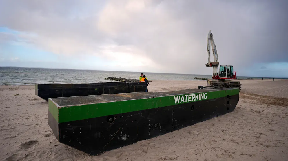Ein Spezial-Schwimmbagger (r.) wird neben Schwimmpontons an den Strand bei Niendorf gefahren, wo ein Wal im flachen Wasser gestrandet ist.