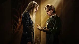 Elrond und Celebrimbor stehen sich im Profil gegenüber und betrachten die leuchtenden Ringe in Celebrimbors Hand. Warmes Licht fällt von oben ein.